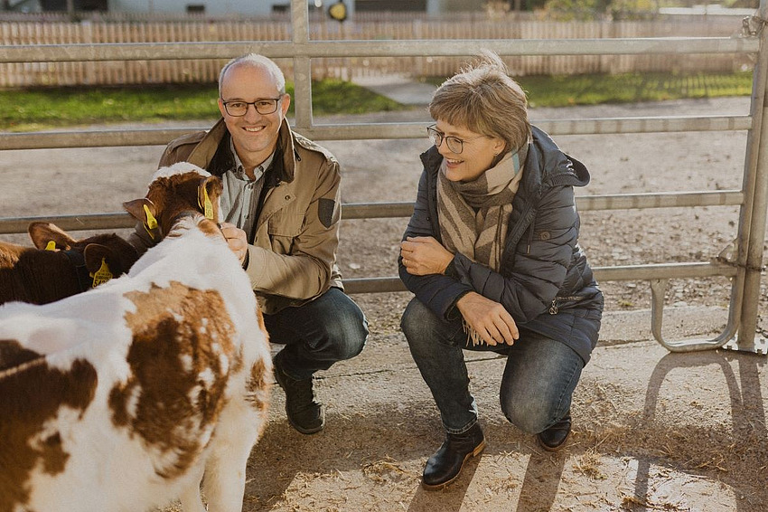 Ein Mann und eine Frau, beide lachend. Der Mann streichelt ein Kalb, welches zu den Personen gedreht ist.